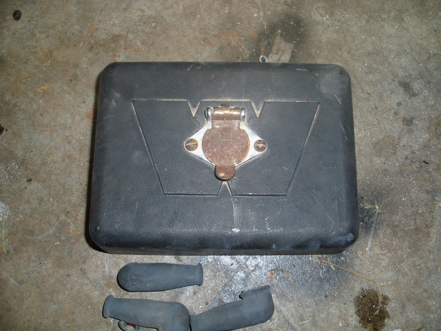 Black rectangular case with metal latch and emblem, on concrete floor with two rubber boots.