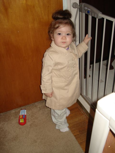 2000 Ford Crown Victoria - A young child wearing a tan trench coat and plaid pants stands next to a baby gate. A toy phone is on the carpet to the left of the child.