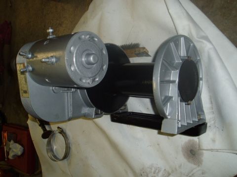 1980 Ford Bronco - A silver and black electric winch is shown resting on a white tarp. The winch has a cylindrical motor housing with bolts and a circular end…