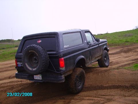 1990 Ford Bronco - A black 1990 Ford Bronco is shown from the rear-right angle, driving on a dirt track with green hills in the background. The vehicle has a…
