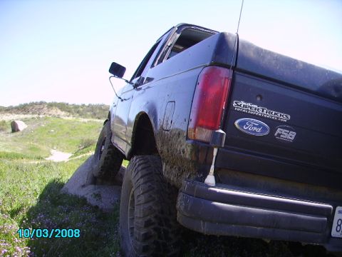 1990 Ford Bronco - A black Ford Bronco is shown from the rear quarter angle, climbing over rocks on a sunny day with green hills in the background. The truck…
