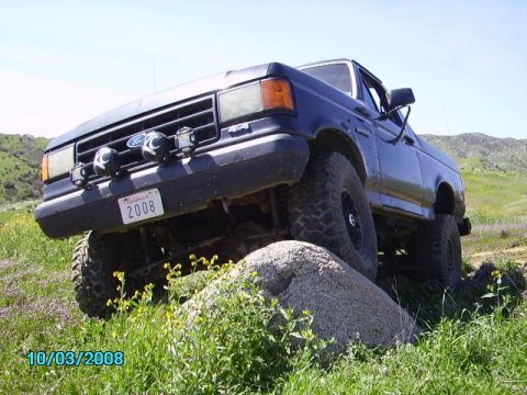 1990 Ford Bronco - A dark blue Ford Bronco is parked on a rocky, grassy hill with purple wildflowers. The front of the vehicle is angled towards the camera, s…