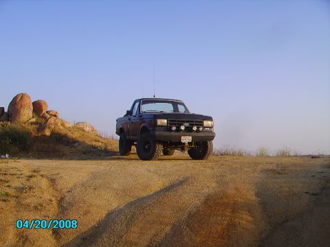 1990 Ford Bronco - A lifted, dark-colored Ford pickup truck with off-road tires is parked on a dirt hill under a clear blue sky. The truck has auxiliary light…