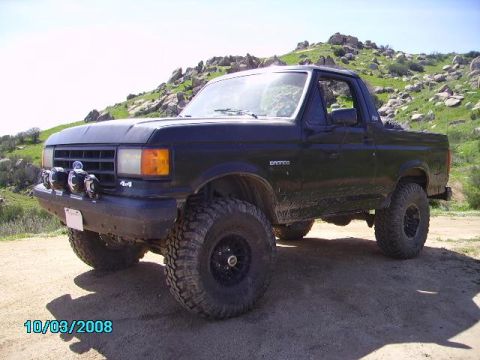 1990 Ford Bronco - A black 1990 Ford Bronco is parked on a dirt road with rocky hills in the background. The vehicle has off-road tires, auxiliary lights on t…