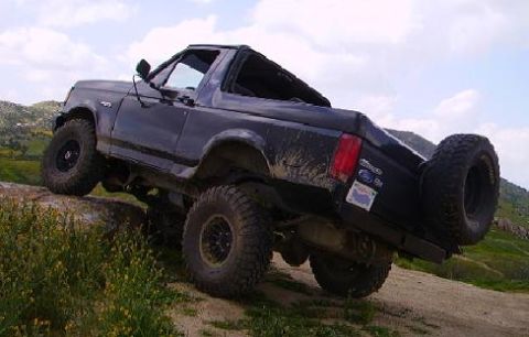 1990 Ford Bronco - A black, lifted Ford Bronco is shown from a rear three-quarter angle, driving off-road over a rocky incline. The vehicle has large, aggress…