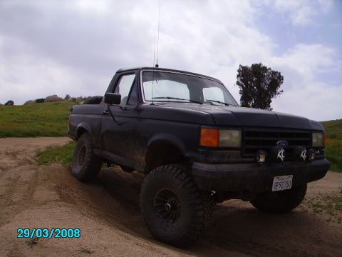 1990 Ford Bronco - A black, lifted 1990 Ford Bronco is parked on a dirt track with grassy hills and a cloudy sky in the background. The vehicle has large off-…
