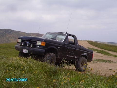1990 Ford Bronco - A black Ford Bronco is parked on a grassy hill next to a dirt road. The vehicle has large off-road tires and auxiliary lights mounted on th…