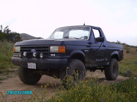 1990 Ford Bronco - A dark blue Ford Bronco is parked on a dirt road in a grassy, hilly area. The vehicle has large off-road tires, KC spotlights mounted on th…
