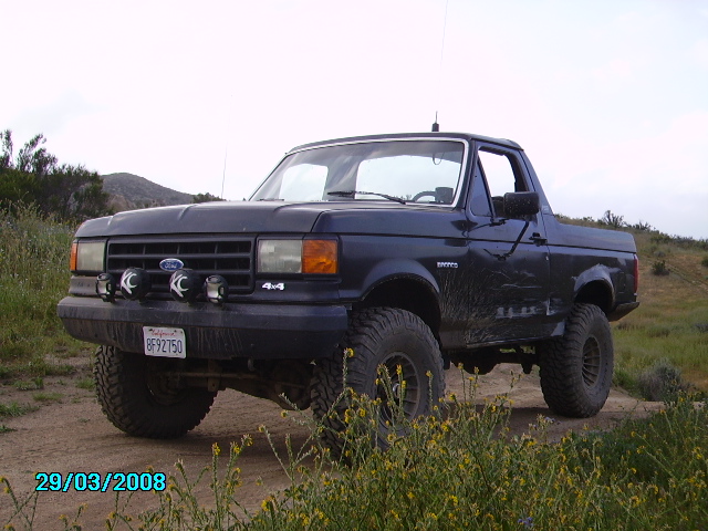 Dark blue Ford Bronco with off-road tires and KC spotlights parked on a dirt road.