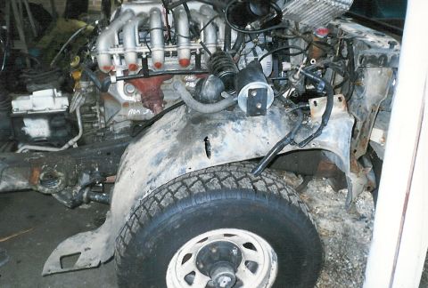 1983 Ford Bronco - The engine and front fender of a heavily damaged vehicle are shown, with a visible tire and wheel.