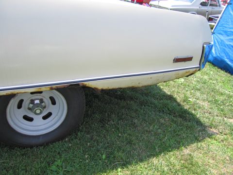 2001 Ford Crown Victoria - Rear quarter panel of a cream-colored vehicle shows rust along the lower body trim and a white steel wheel.