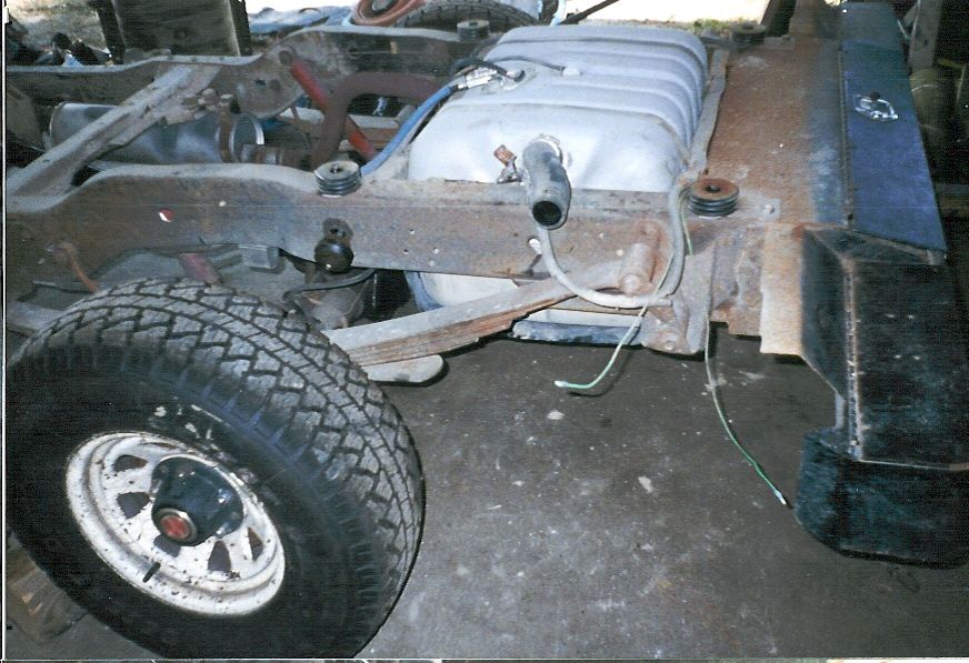 Bare rusty truck chassis with mounted wheel and silver fuel tank on dirt floor