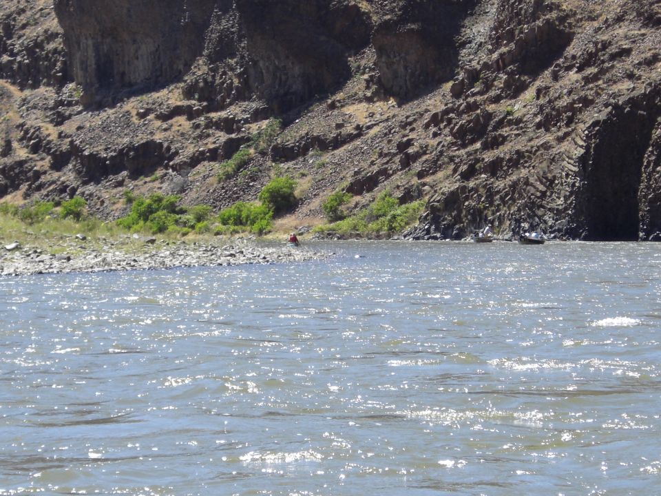 River flows past a rocky cliff with sparse vegetation; two small boats are visible near the shore