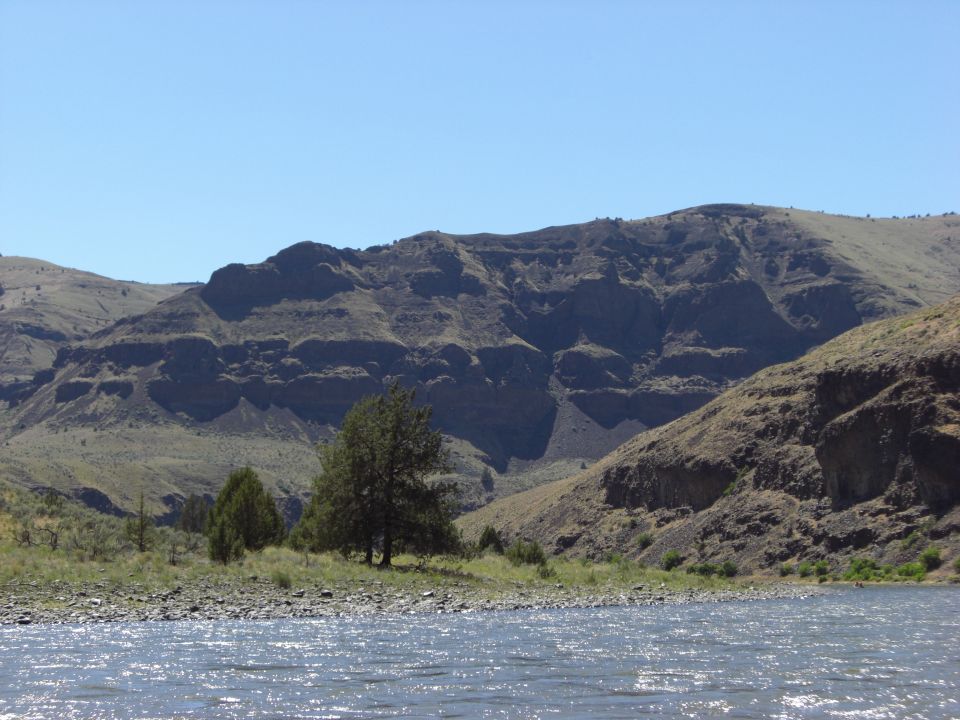 River flows past rocky shore with trees and layered mountains under a clear blue sky