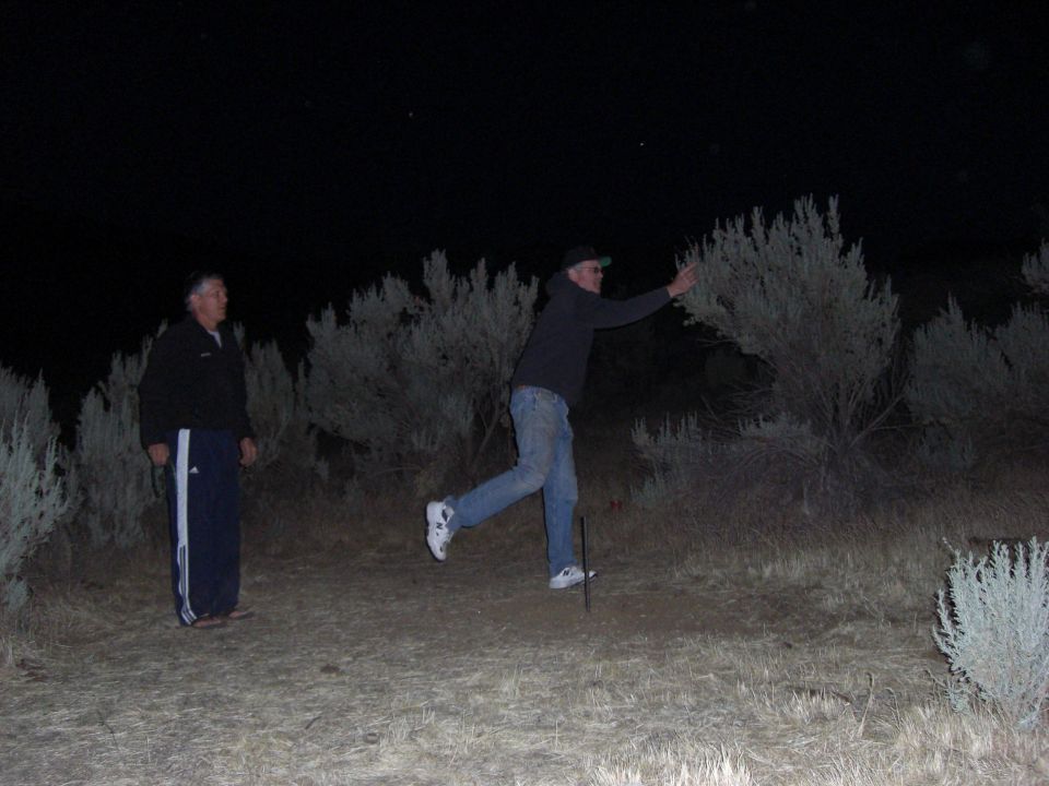 Two men stand in a dry, brushy field at night under a starry sky. One man in a dark jacket and blue pants looks to the left. The other man in a hoodie and jeans is in motion, reaching out to the right