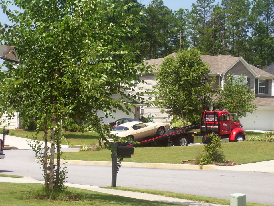 Cream-colored coupe on a red tow truck, viewed from the front-right. The car has a long hood and a sloping rear. A second dark car is parked behind it