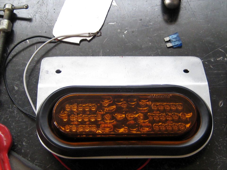 Oval amber LED light with "Maxxima" branding, mounted on a metal plate with wires and a fuse nearby