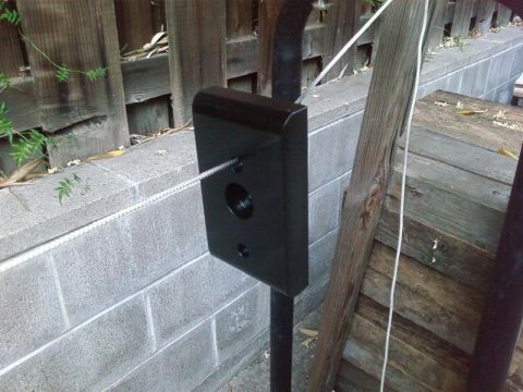 1995 Ford Bronco - A black metal component with a cable running through it is mounted on a pole, with a cinder block wall and wooden fence in the background.