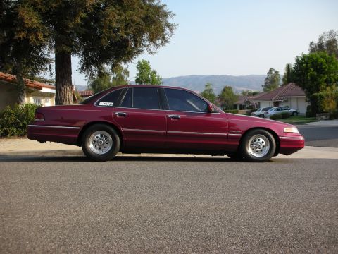 1992 Ford Crown Victoria - Full profile view of a maroon Ford Crown Victoria sedan parked on a suburban street with tile-roofed houses and hills behind. It sits on po…