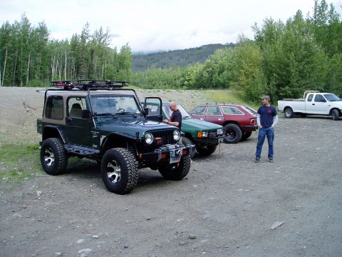 2000 Ford E350 - A dark green Jeep Wrangler is parked on a gravel lot with a white Ford pickup truck in the background. A man in a blue shirt and jeans stan…