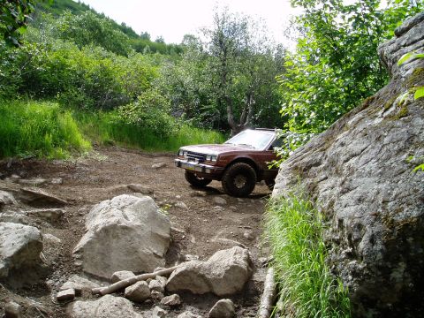 2000 Ford E350 - A maroon 1980s sedan is parked on a dirt path surrounded by lush green vegetation and large rocks. The vehicle is angled towards the right,…