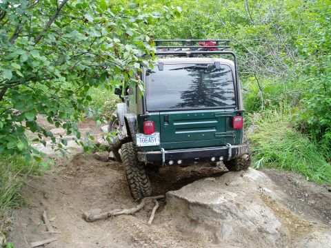 2000 Ford E350 - A dark green Jeep Wrangler is navigating a steep, rocky, and muddy off-road trail. The rear of the vehicle is visible, showing its tires, b…