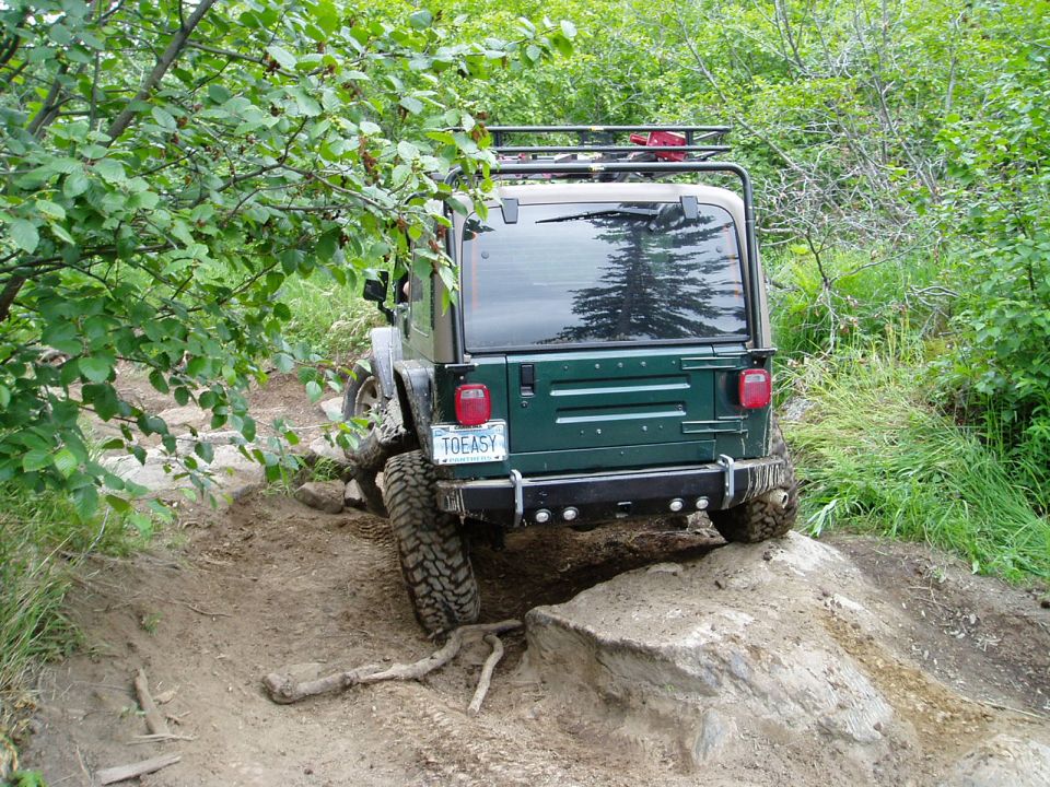Green Jeep Wrangler on a muddy, rocky off-road trail, rear view.