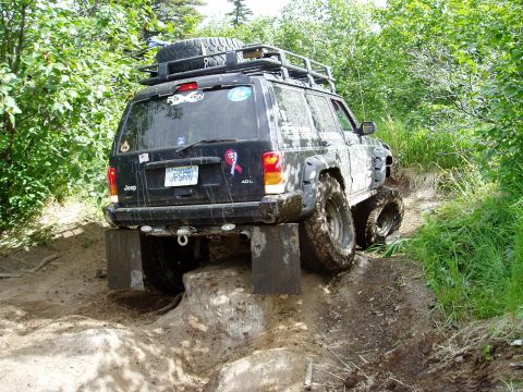2000 Ford E350 - A black Jeep Cherokee is navigating an off-road trail, with its rear wheels elevated on blocks and the front wheels in a muddy rut. The veh…