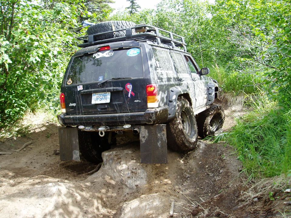 Black Jeep Cherokee off-roading, rear wheels on blocks, front in mud, surrounded by trees.