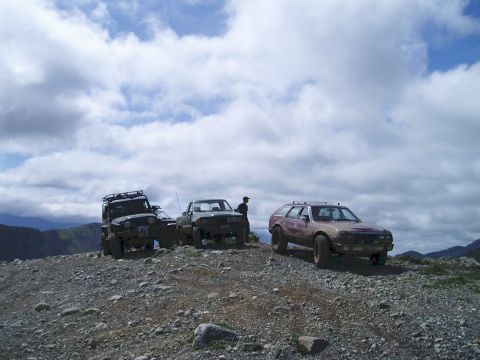 2000 Ford E350 - Three off-road vehicles are parked on a rocky, mountainous terrain under a cloudy sky. A person stands next to the rightmost vehicle, a tan…
