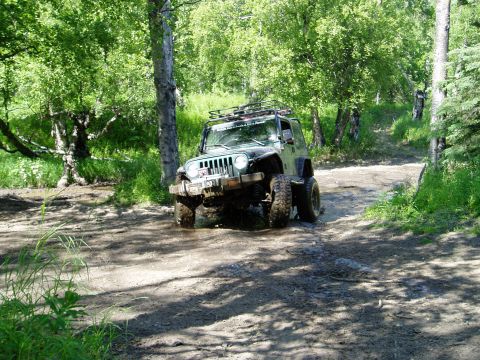 2000 Ford E350 - A green Jeep Wrangler is driving through a muddy, wooded trail. The vehicle is angled towards the viewer, with its front tires deep in the…