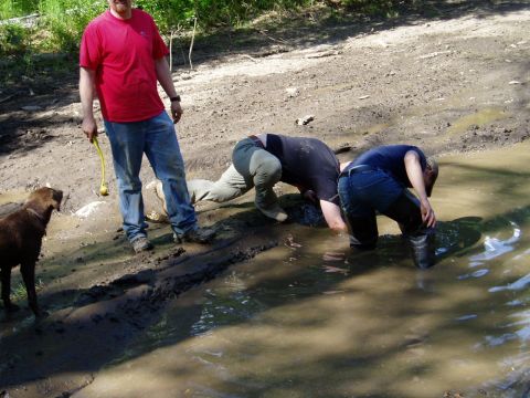 2000 Ford E350 - A man in a red shirt stands next to a brown dog, holding a yellow toy. Two people are bent over in a muddy puddle in the background.