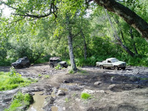 2000 Ford E350 - Two off-road SUVs and a pickup truck are navigating a muddy, wooded trail with puddles and rough terrain. The vehicles appear to be in moti…