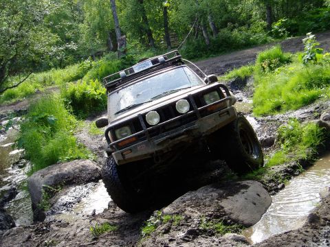 2000 Ford E350 - A tan 4x4 SUV is navigating a muddy, rocky trail with water obstacles, its front tires angled to the right as it climbs over a rock.