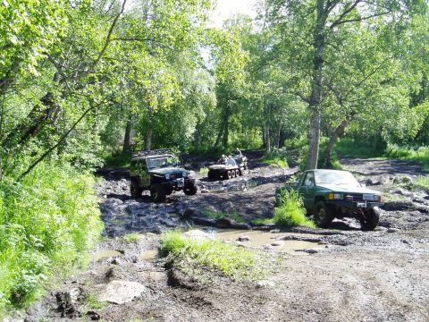 2000 Ford E350 - Two off-road vehicles, a green Jeep Wrangler and a green pickup truck, are navigating a muddy trail surrounded by lush green trees. Another…