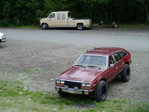 2000 Ford E350 - A lifted, maroon AMC Eagle wagon with large off-road tires is parked on a gravel lot. In the background, a tan Ford E-Series van cutaway ch…
