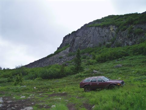 2000 Ford E350 - A maroon station wagon with a roof rack is parked on a grassy hillside below a rocky cliff face. The vehicle is angled towards the right, w…