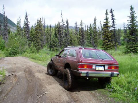 2000 Ford E350 - A maroon station wagon with off-road tires is parked on a dirt road in a wooded area with tall evergreen trees.