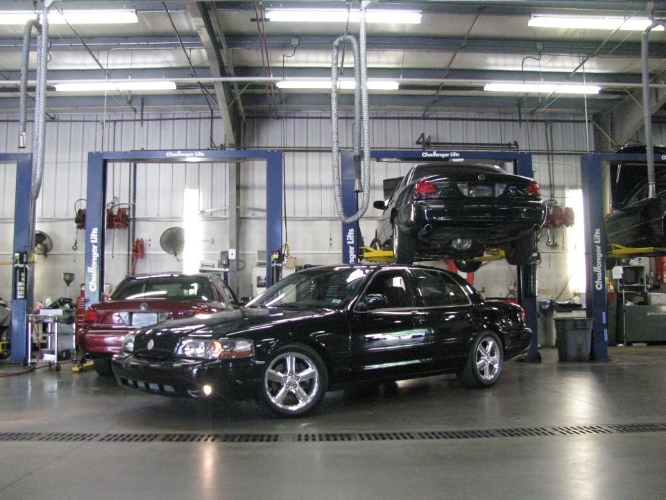 Two sedans in a garage, one black in the foreground, one red behind it, and another black sedan on a lift.