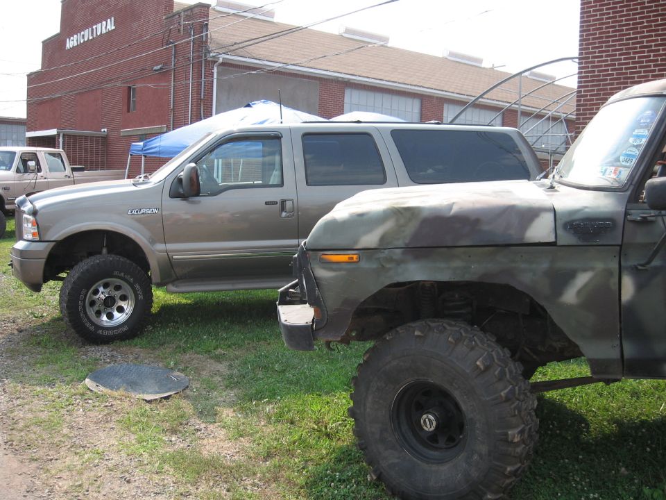 Tan Ford Excursion SUV parked next to a camouflage truck. Agricultural building in background.