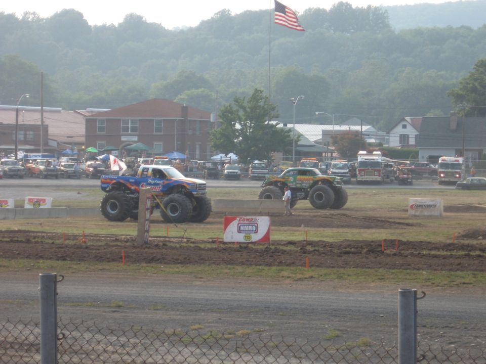 Two monster trucks, one blue and white, the other green and yellow, on a dirt track.