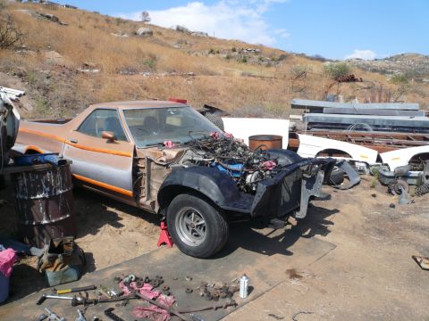 1975 Ford Ranchero - A partially disassembled orange and silver Ford Ranchero sits in a junkyard setting with various car parts scattered around it.