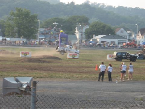 1984 Ford Bronco - A truck soars through the air during an event, with spectators and another vehicle watching.