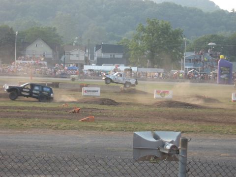1984 Ford Bronco - Trucks compete in a dirt race, with one vehicle mid-air over a jump and the other navigating the track below.
