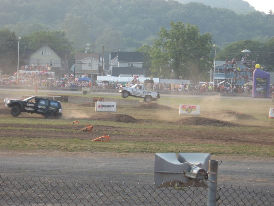 Two trucks in a dirt race, one airborne over a mound, the other on the ground kicking up dust