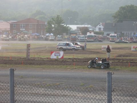 1984 Ford Bronco - A blue and white 1984 Ford Bronco is seen on a dirt track alongside other vehicles and spectators.
