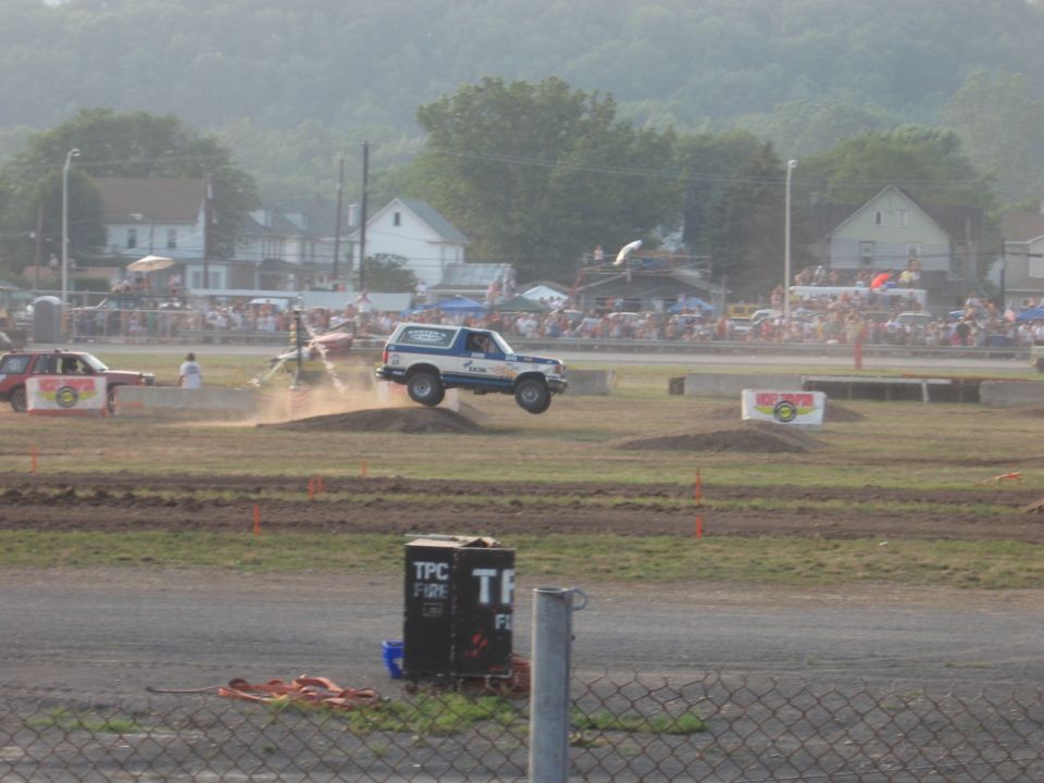 Ford Bronco airborne over dirt jump, another car on left, crowd in background