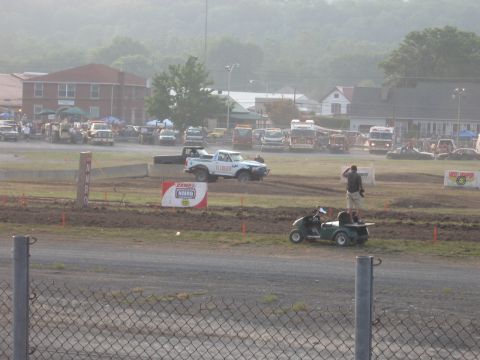 1984 Ford Bronco - A white pickup truck navigates a muddy track at an outdoor event with spectators in the background.