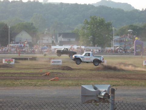 1984 Ford Bronco - Two pickup trucks catch air during a race event, with spectators watching from the stands.