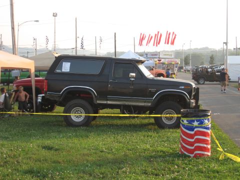 1984 Ford Bronco - A black Ford Bronco is parked on a grassy area with a crowd and vendor tents in the background.